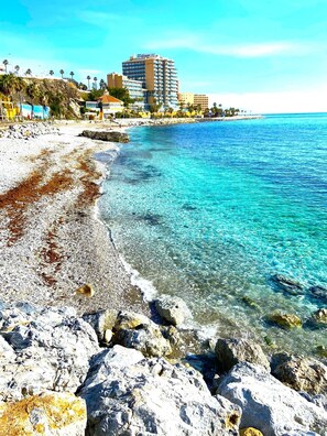 A beach with clear turquoise water, rocky shoreline, and a hotel building in the background. - Aloha Playa Deluxe: Wi-Fi, Swimming Pools, and Parking (Benalmádena)
