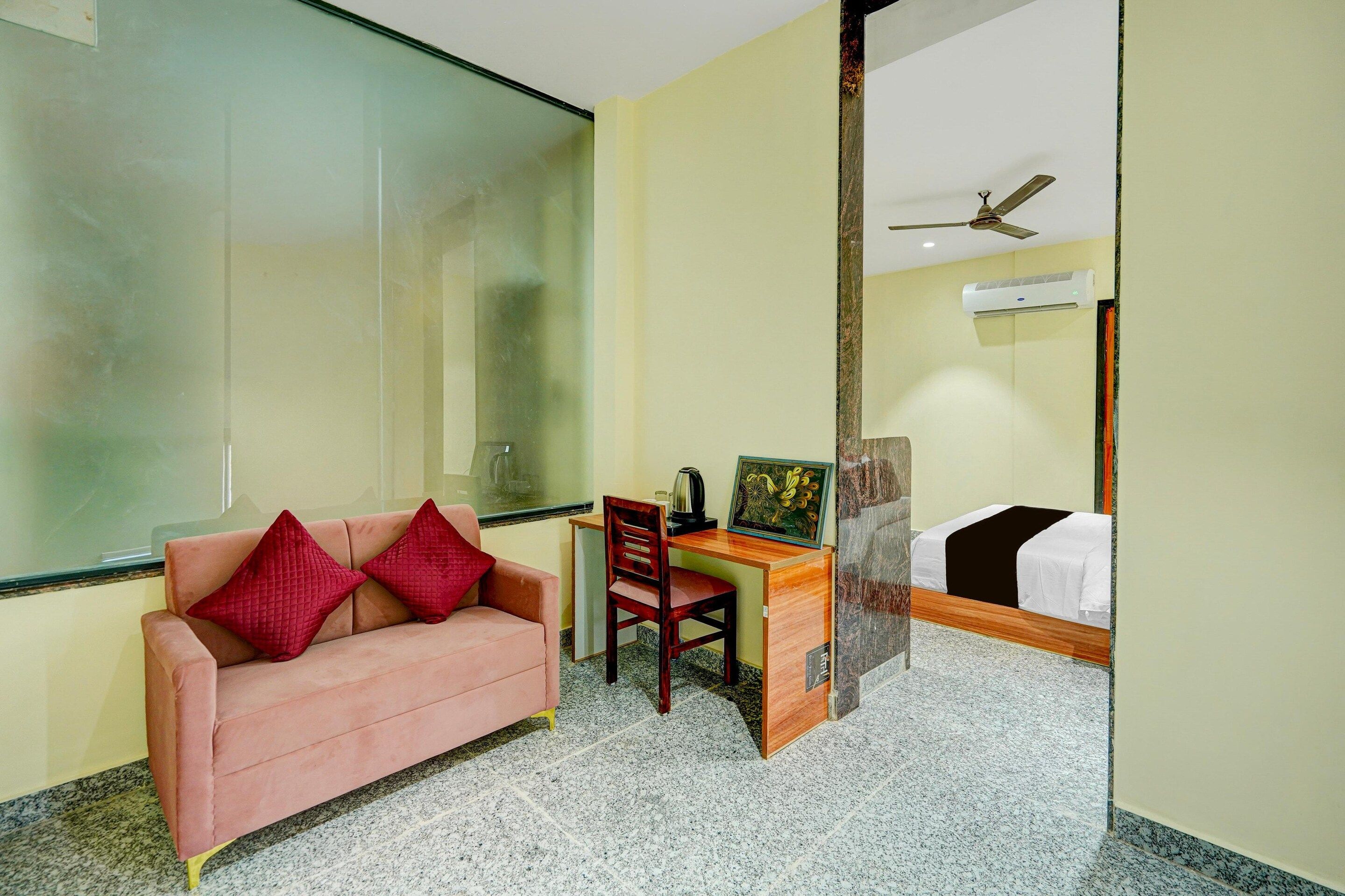 A hotel room with a pink sofa, a wooden desk, a bed, a ceiling fan, and a large window.