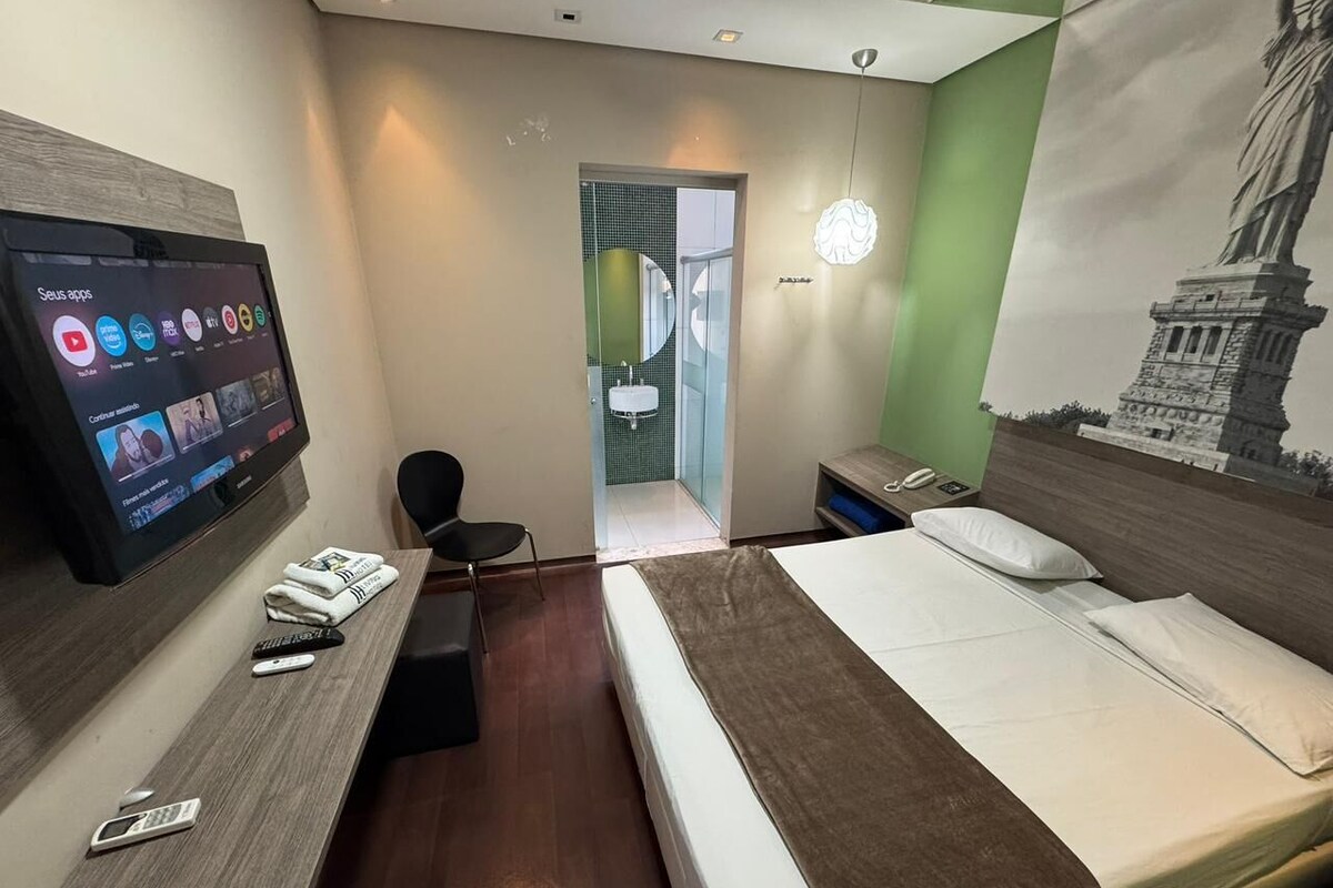 A hotel room with a bed, a desk with a TV, a chair, and a bathroom visible through an open door.