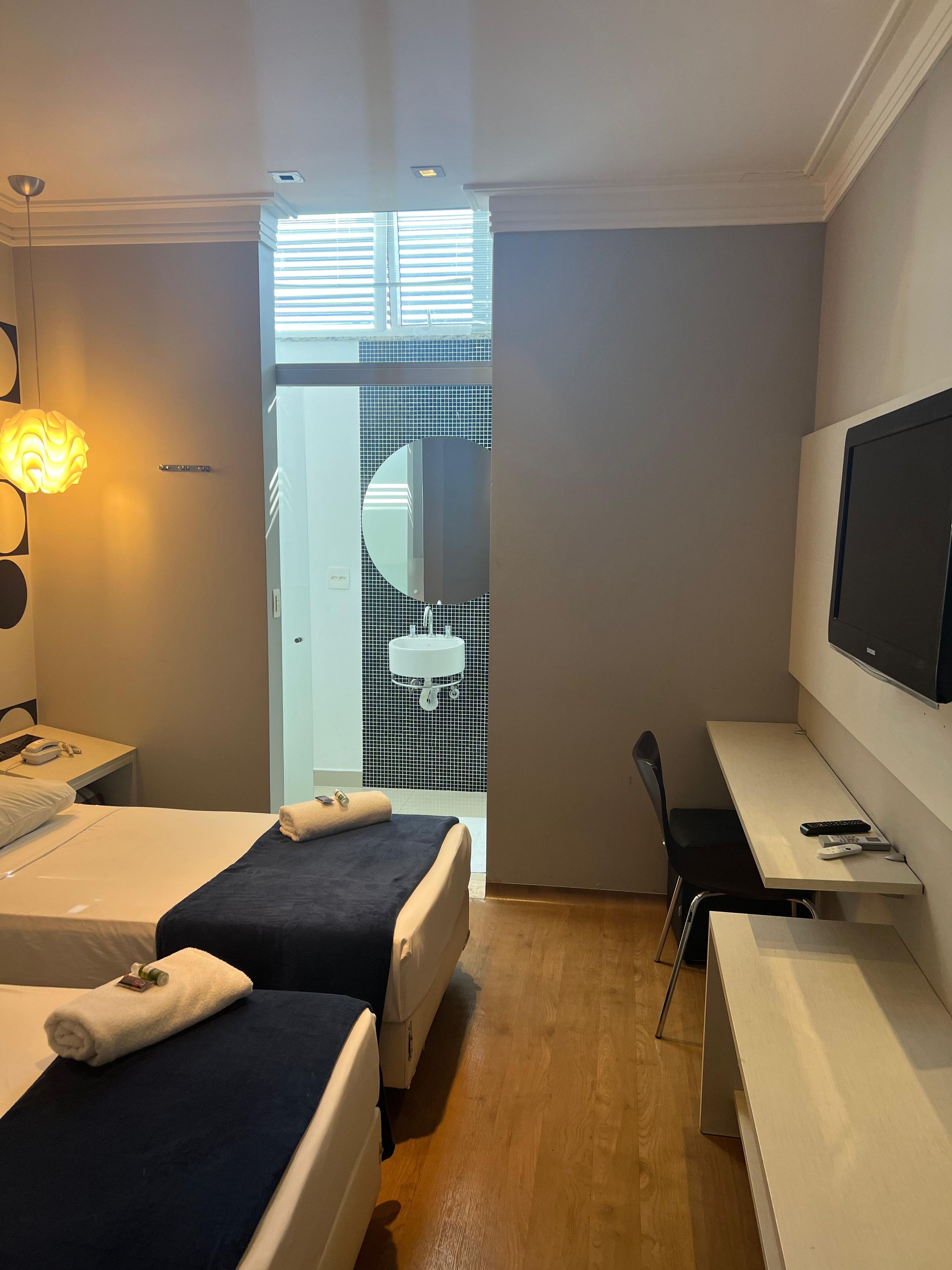 Ein Hotelzimmer mit zwei Betten, einem Schreibtisch, einem Sessel, einem Fernseher und einem Badezimmer mit Spiegel und Waschbecken.