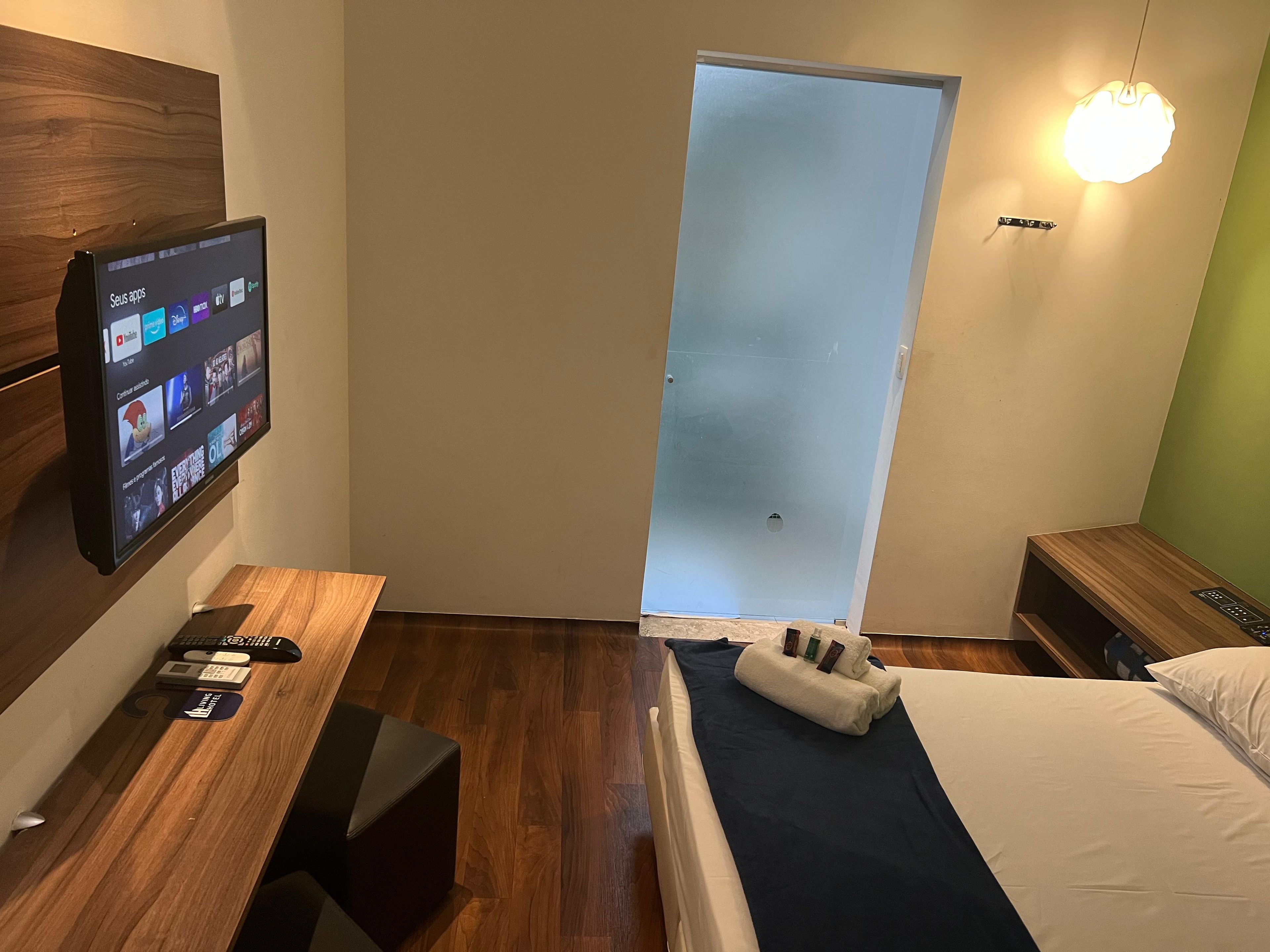 Habitación de hotel con cama, escritorio con monitor de computadora que muestra aplicaciones, mesita de noche y lámpara de pared.