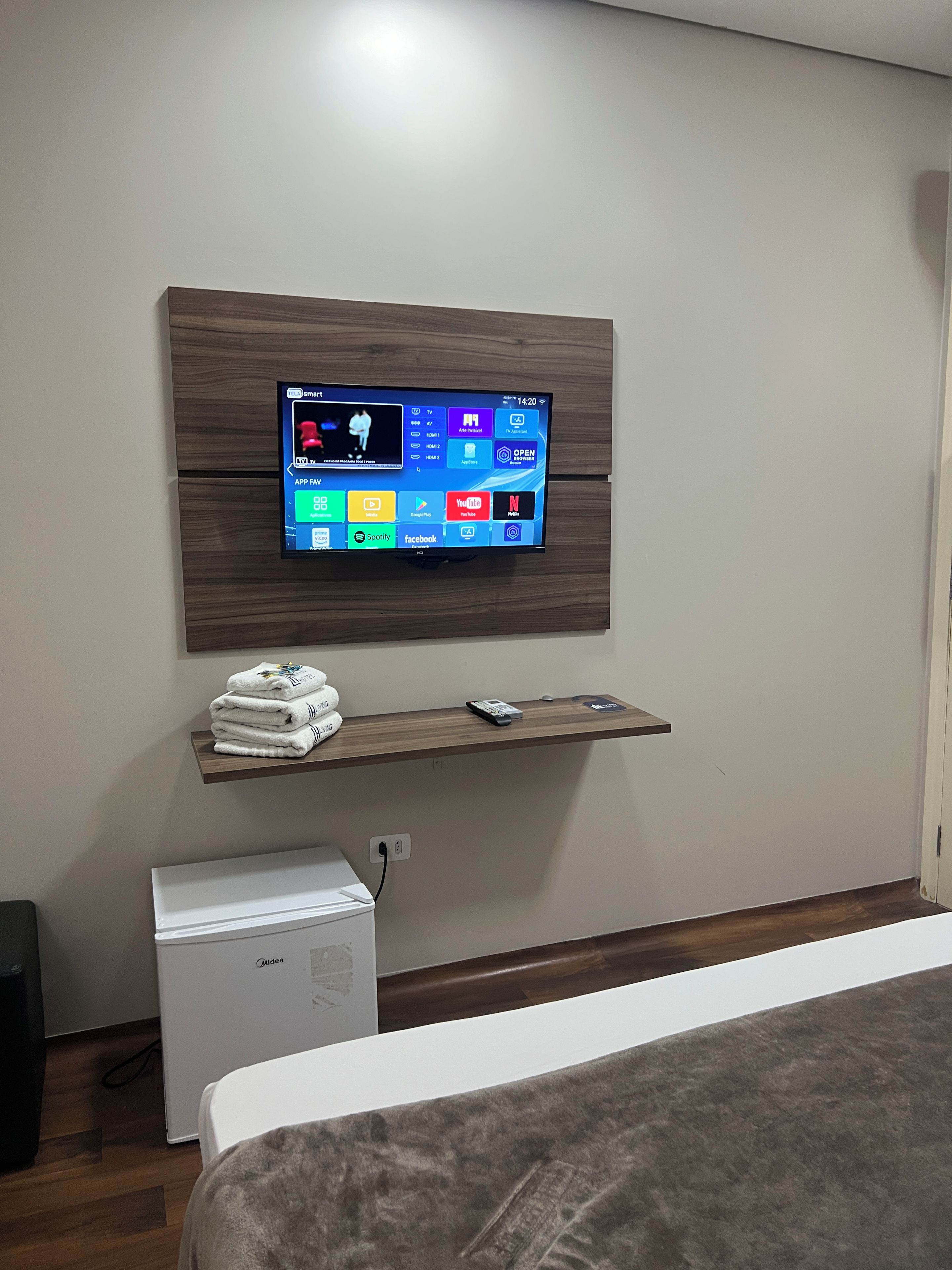 Un televisor montado en una pared de madera, con un estante debajo que muestra un control remoto y una pila de toallas.