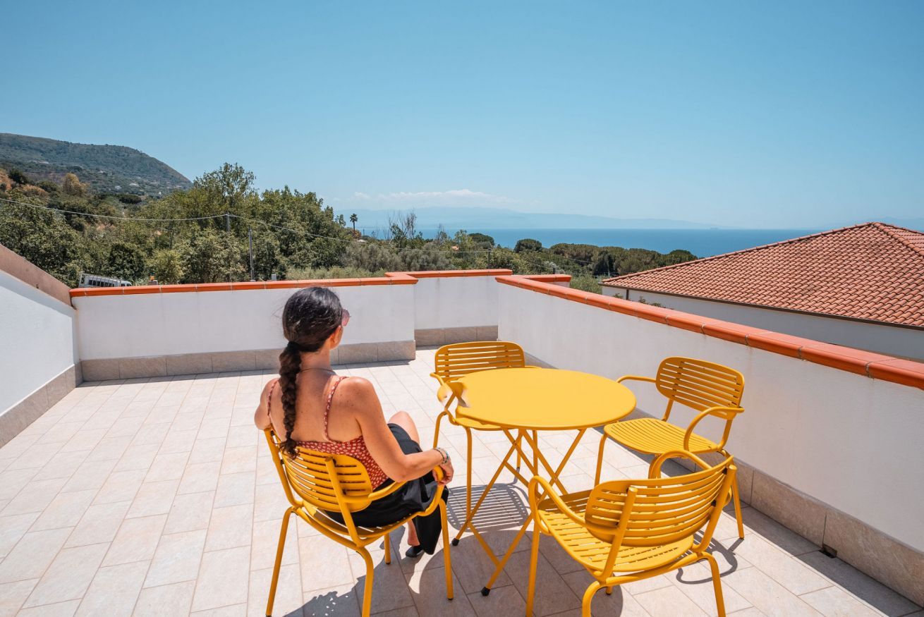 Una persona sentada en una mesa y sillas de metal amarillo en un balcón, con vista al mar y las colinas.