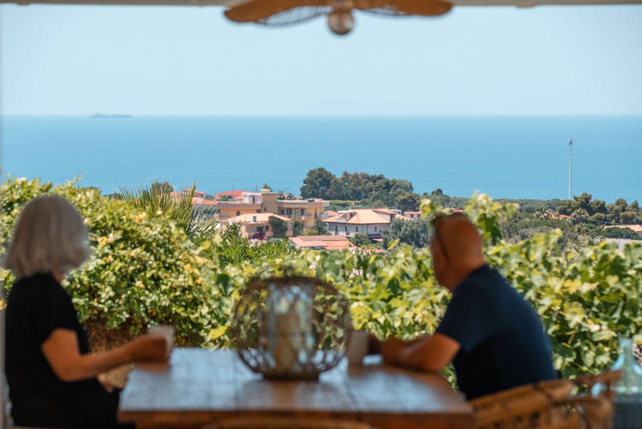 Un couple est assis à une table, avec vue sur la mer et sur des bâtiments qui se dessinent à l'horizon.