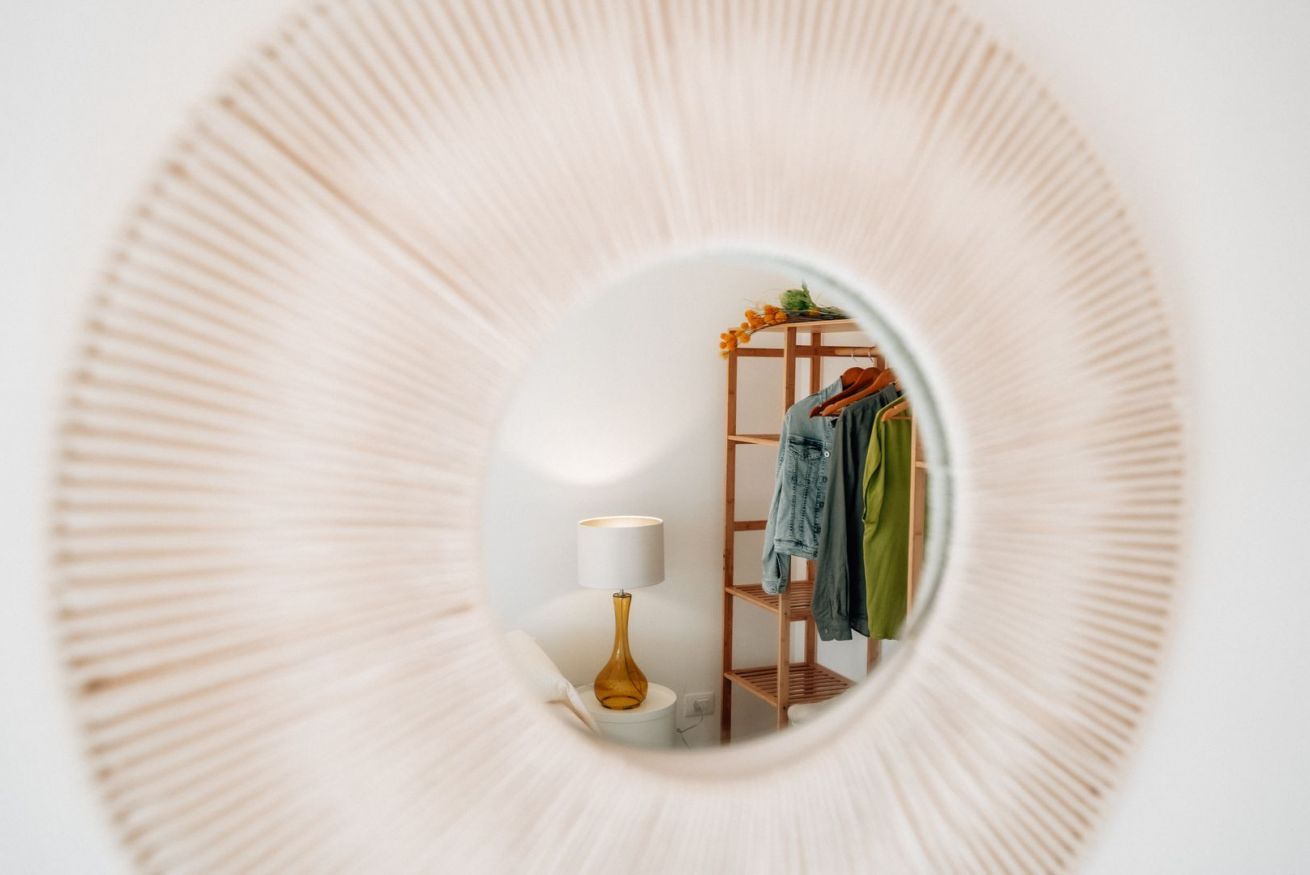 Un miroir rond reflète une pièce dans laquelle on aperçoit une étagère en bois, une lampe et des vêtements suspendus.