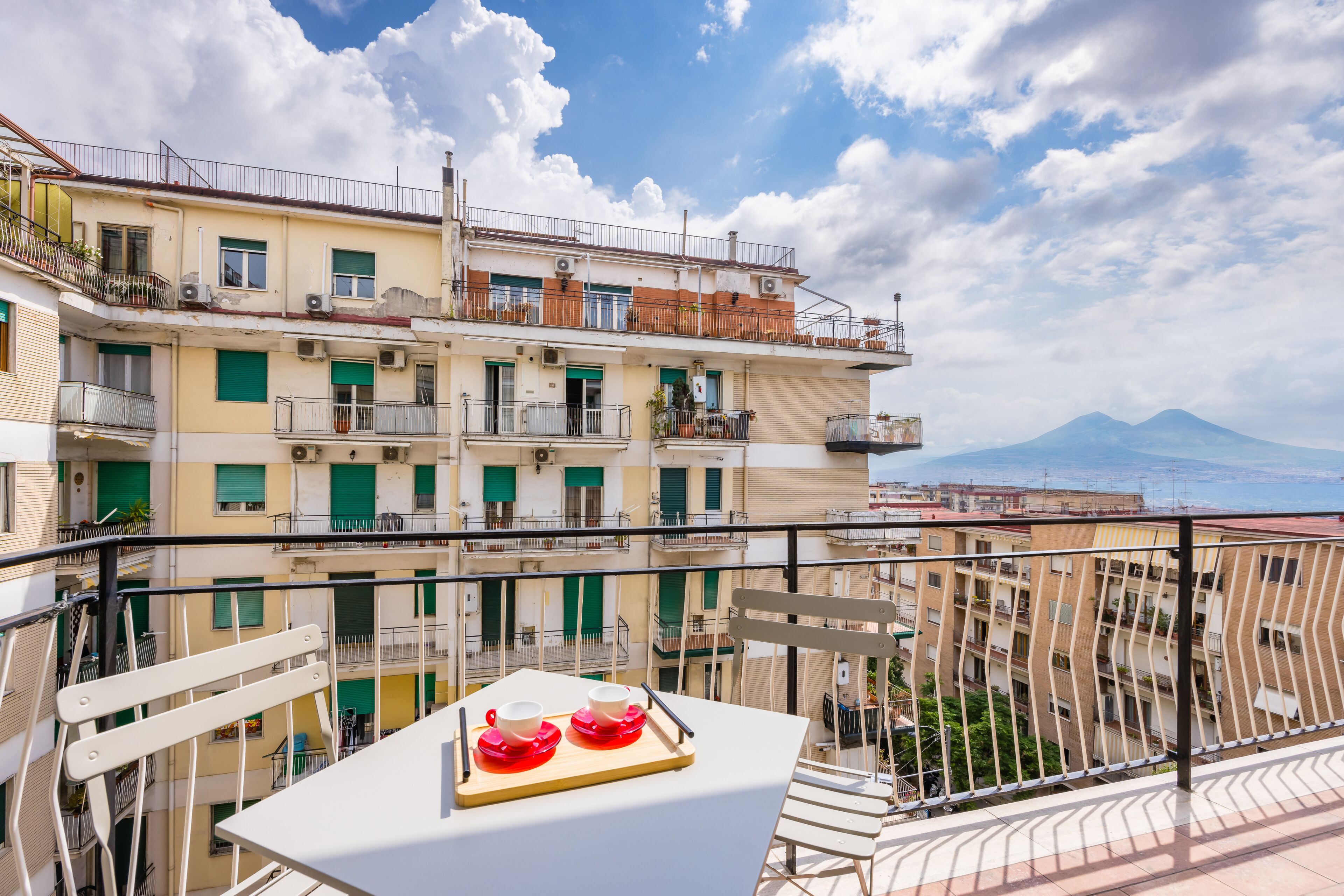 A balcony with a table set for two, overlooking a building with multiple balconies and a mountain in the background.