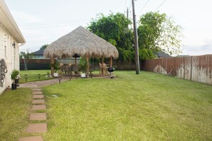 A backyard with a thatched-roof gazebo, a barbecue grill, and a paved pathway leading to the house.