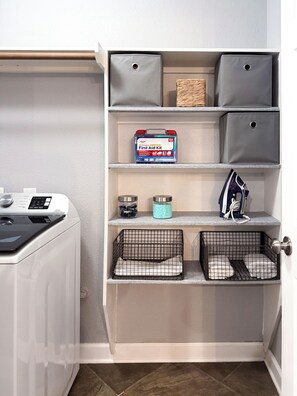 A laundry area with a washing machine, shelves with storage bins, and a first aid kit. - Your home away from home at Casa Palapa (Missouri City)