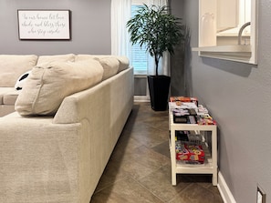 A living room with a beige sofa, a potted plant, a white side table with board games, and a framed quote on the wall. - Your home away from home at Casa Palapa (Missouri City)