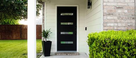 A black front door with a small window, a white porch, a brick wall, and a well-maintained hedge.