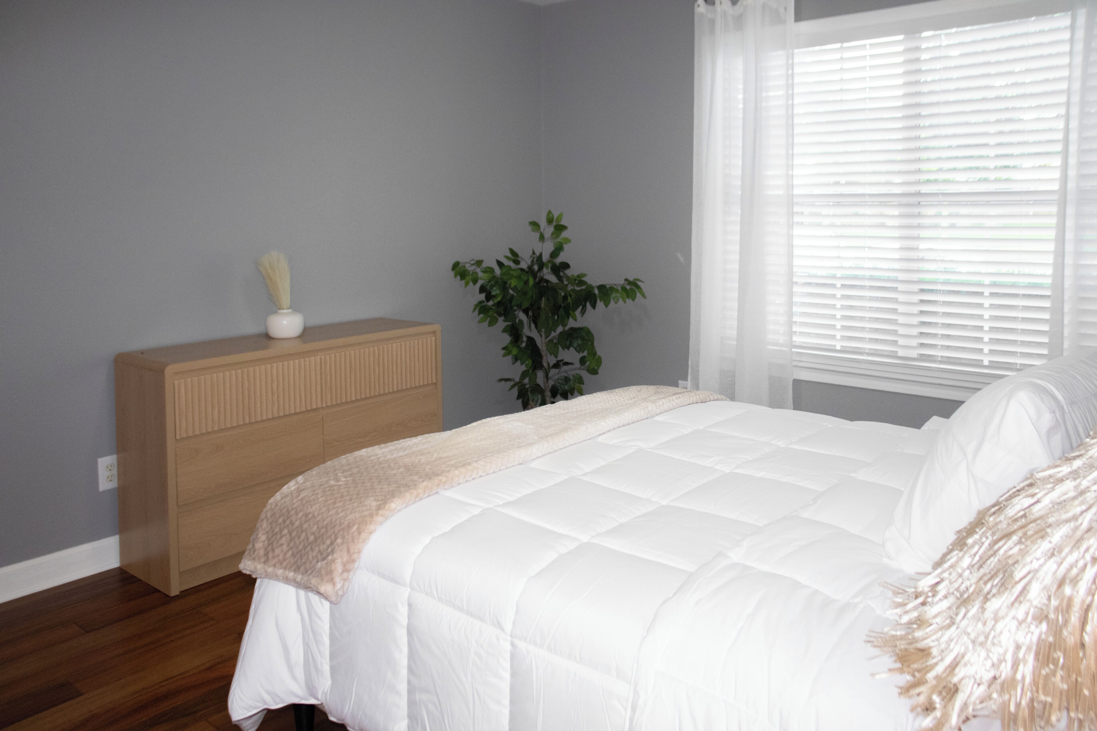 Een slaapkamer met een bed, een dressoir, een plant en een raam met jaloezieën.