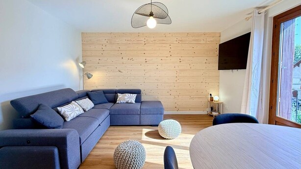 A modern living room with a sectional sofa, round ottomans, a dining table, and a wooden wall. - 'Le Belle Vue' Apartment: Mountain View, Shared Garden & Wi-Fi (Villard-de-Lans)