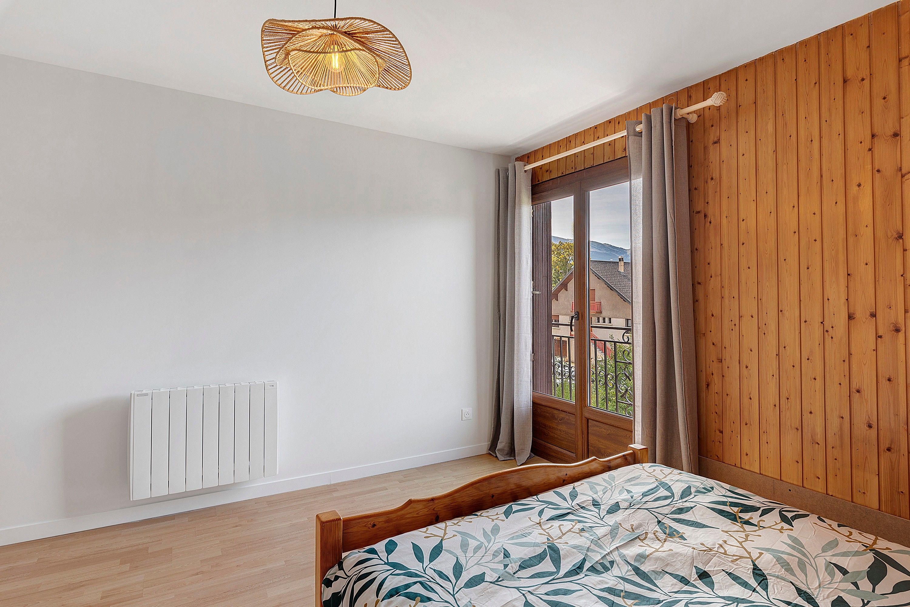 Un dormitorio con cama, radiador, pared de madera, ventana con cortinas y lámpara colgante.