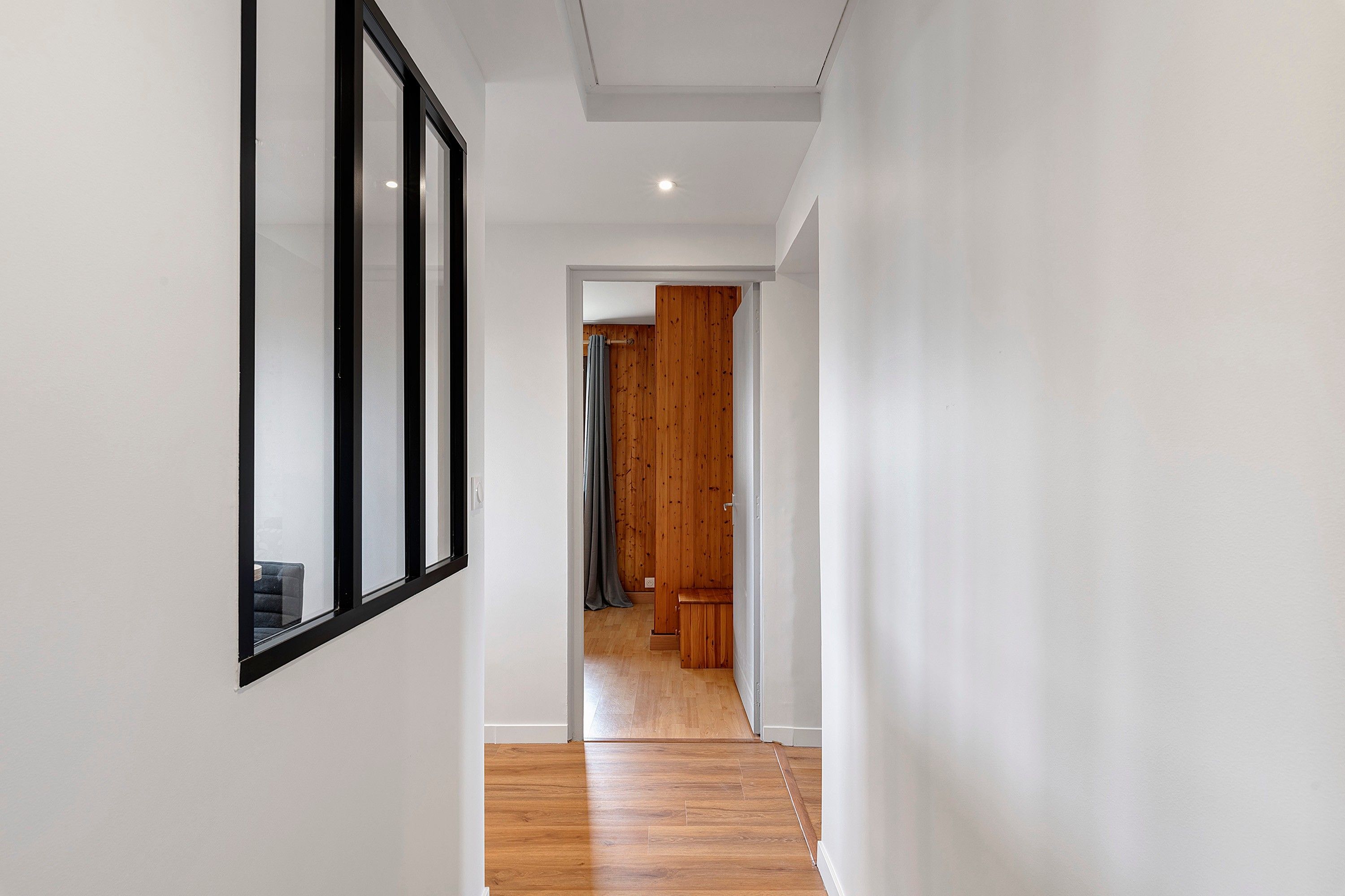 Un pasillo con piso de madera, paredes blancas y una puerta que da a otra habitación.