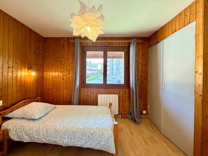 Una habitación con paredes de madera, una cama con colcha estampada, una ventana con vista a edificios y una lámpara en el techo.