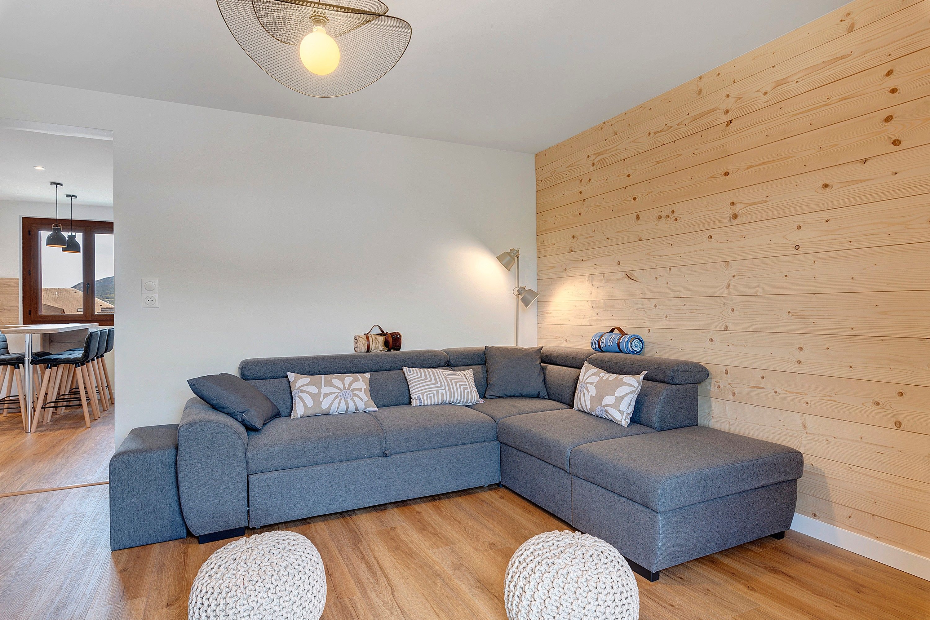 A modern living room with a grey sectional sofa, wooden wall paneling, a round knitted rug, and a pendant light.