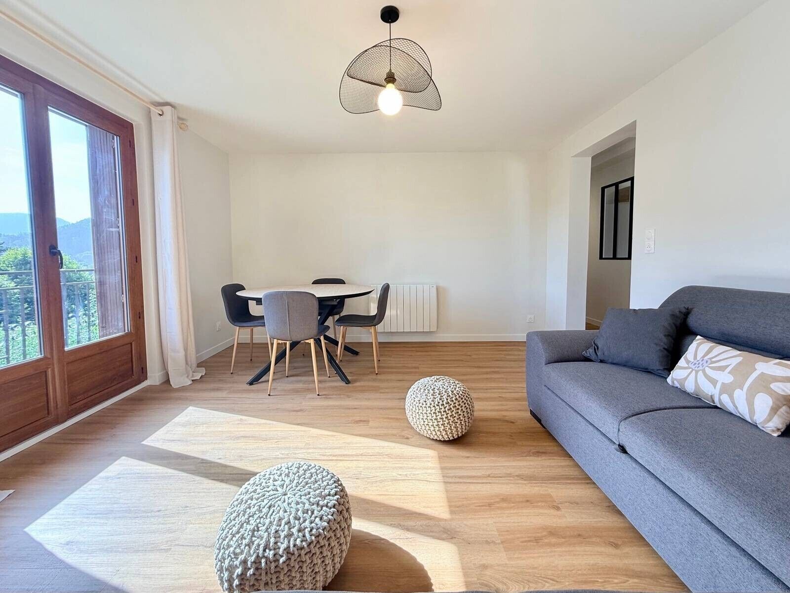 Una sala moderna con un sofá gris, un otoman redondo y una zona de comedor con mesa y sillas.