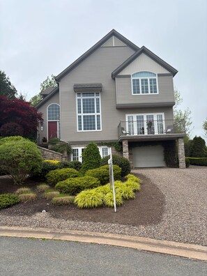 A two-story house with a garage, a driveway, and a well-maintained garden. - Elegant 4BR Family Home-Hot Tub, Backyard Trails & Reservoir Access (30 days) (Wolfville)