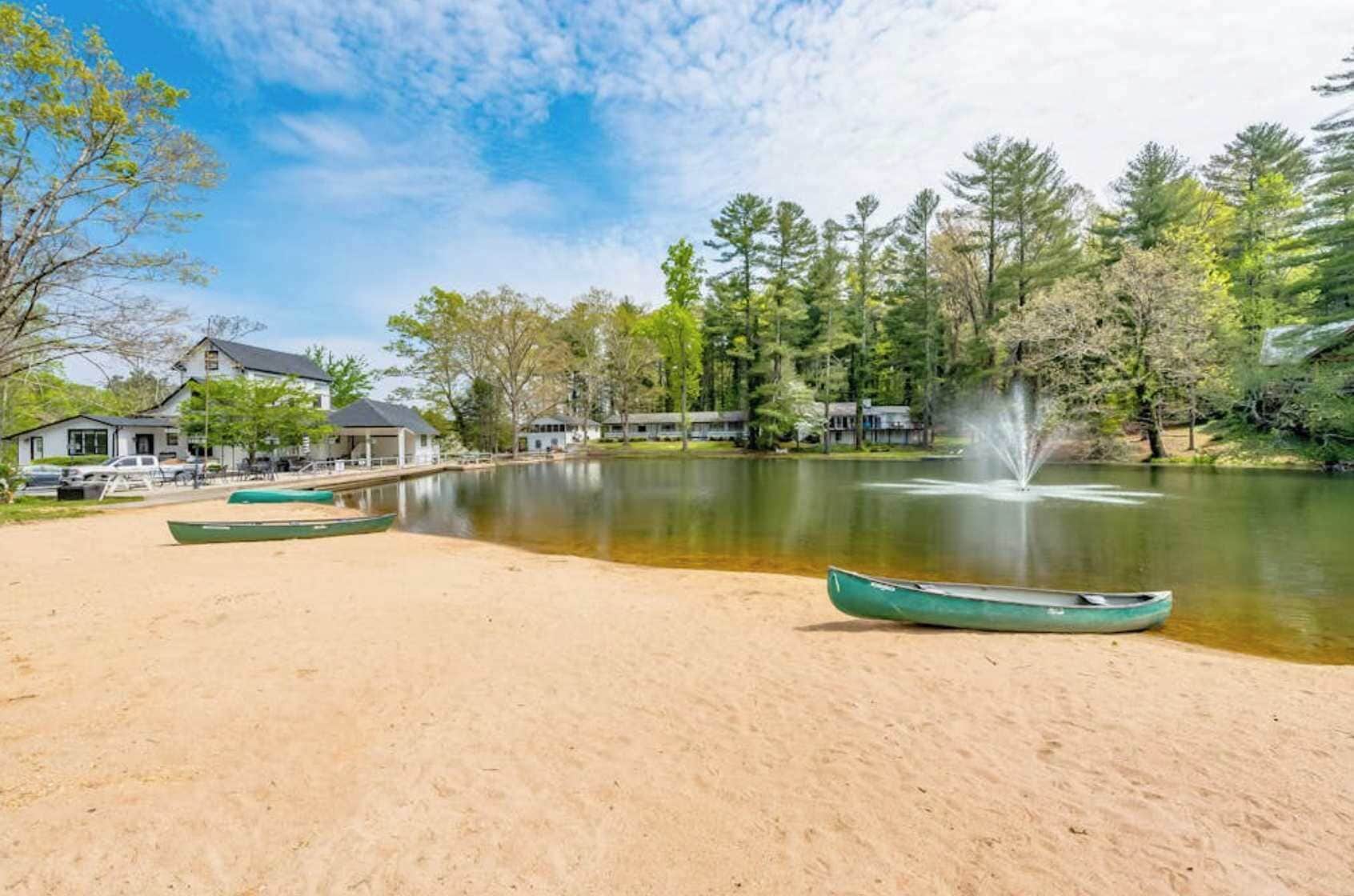 Un entorno junto al lago con playa de arena, canoas verdes, una fuente y casas al fondo.