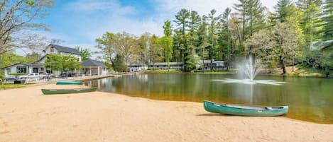 A lakeside setting with a sandy beach, green canoes, a fountain, and houses in the background.