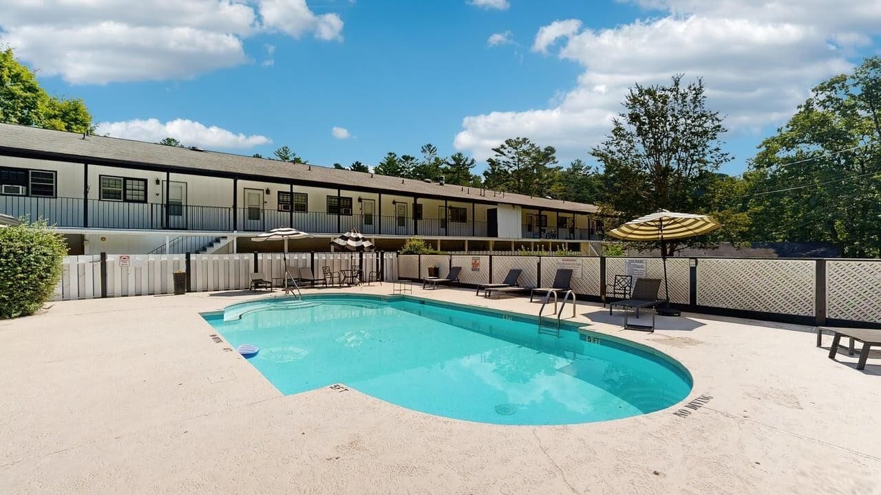 Piscina com espreguiçadeiras e guarda-sóis, cercada por uma área vedada e um edifício ao fundo.