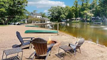 A lakeside setting with a fire pit, chairs, and canoes by the water.