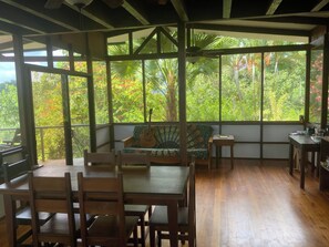 Une pièce avec une table et des chaises en bois, un canapé recouvert d’un tissu à motifs et de grandes fenêtres donnant sur une végétation tropicale.