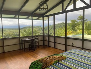 Une chambre avec un lit, un bureau et une chaise, de grandes fenêtres donnant sur un paysage boisé.