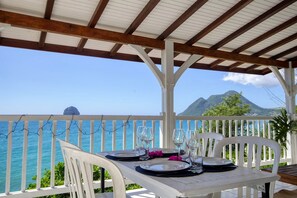 A covered outdoor dining area with a table set for two, overlooking a coastal view with a mountain in the distance. - Vrbo Property (Le Diamant)
