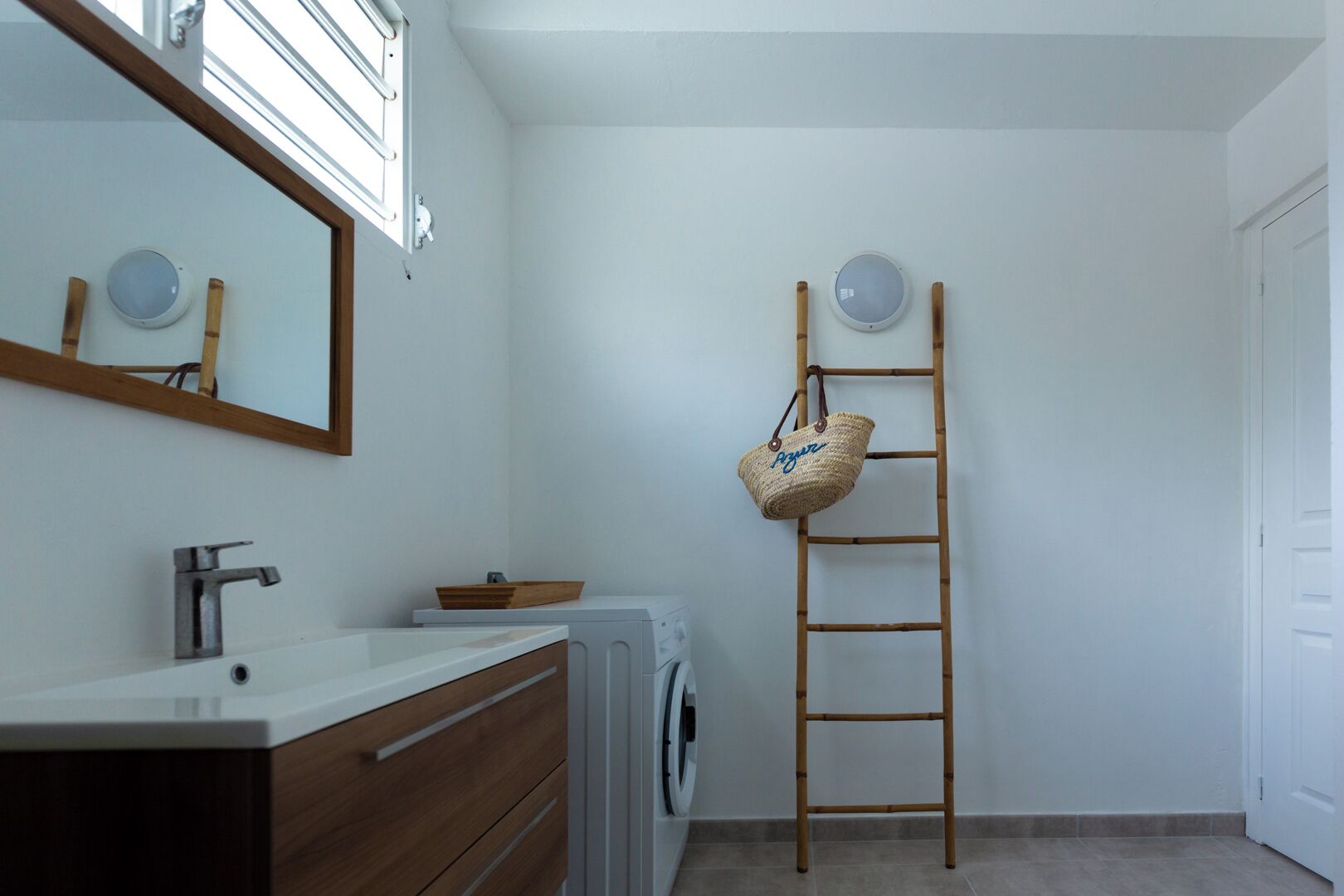 A bathroom with a wooden vanity, a washing machine, a mirror, and a wooden ladder with a woven bag.