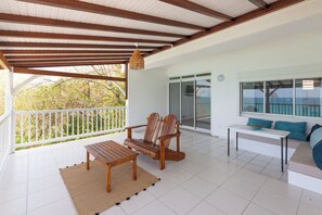 A covered outdoor area with a wooden table, two chairs, a sofa, and a hanging light fixture. - At Alexandra's, AZUR Apartment (Le Diamant)