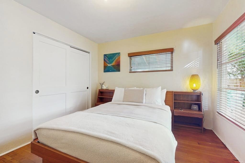A bedroom with a bed, a nightstand, a lamp, a window with blinds, and a door.