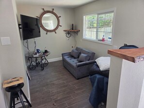 A compact living space with a grey sofa, a television mounted on the wall, a wooden shelf, and a window with a view of the outside. - Fully Remodeled and Updated, just 90-mins from San Francisco! (Monte Rio)
