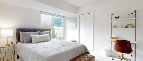 A bedroom with a large bed, a bench, a desk with a chair, and a shelving unit.