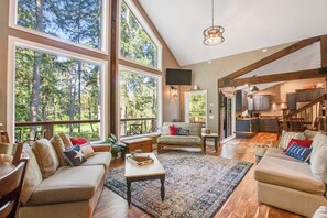 A spacious living area with large windows, a cozy sofa set, a coffee table, and a kitchen in the background.