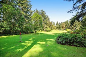 A well-maintained grassy area with a disc golf basket, surrounded by dense trees and a clear sky. - Welcoming 2BR Lakeview - Deck - Firepit - Washer Dryer (Anderson Island)