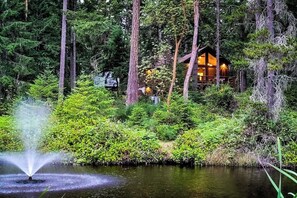 A cabin surrounded by dense forest, a pond with a fountain, and a clear view of the cabin's interior. - Welcoming 2BR Lakeview - Deck - Firepit - Washer Dryer (Anderson Island)