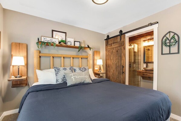Un dormitorio con una cama grande, cabecera de madera, lámparas de noche, un armario con puerta corrediza tipo granero, y un baño con lavamanos y espejo.