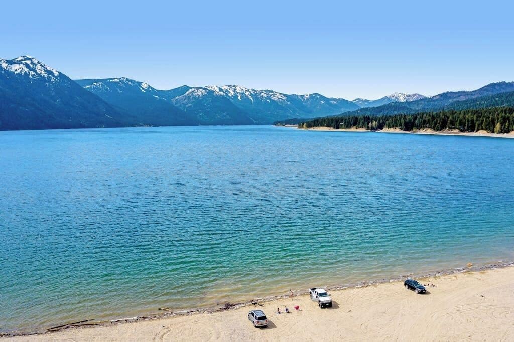 Ein See mit Bergen im Hintergrund und einem Sandstrand, auf dem Fahrzeuge stehen.