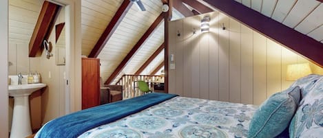 Un dormitorio con techo inclinado, una cama con ropa de cama estampada, un ventilador de techo y una mesita de noche con lámpara.