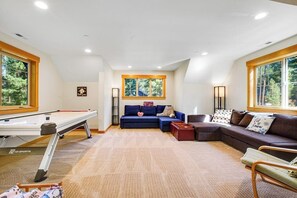 Quarto com mesa de bilhar, um sofá azul, um sofá marrom e janela com vista para árvores.