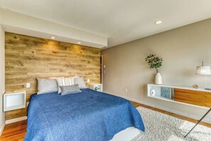 A bedroom with a wooden headboard, a bed with a blue bedspread, a white nightstand, a bedside table, and a wall-mounted shelf with decorative items. - 4BR Bayview Dog Friendly - Balcony - Firepit (Brinnon)