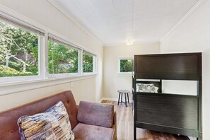 Ein Zimmer mit einer Couch, einem kleinen Hocker und einem dunklen Bücherregal. Fenster sorgen für natürliches Licht und einen Blick ins Grüne.