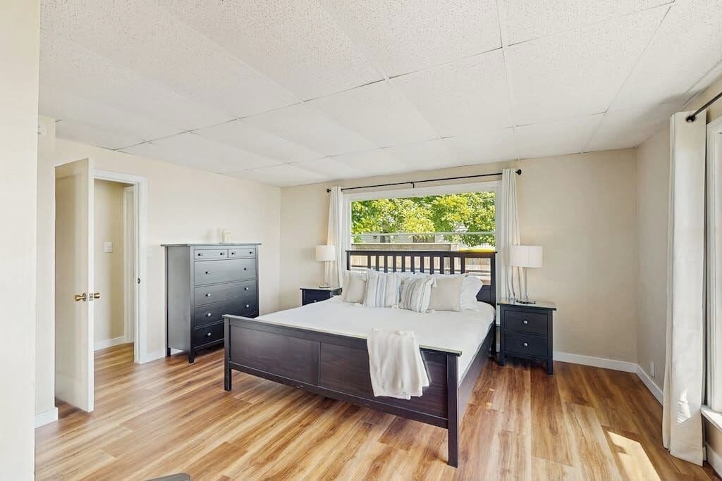 A bedroom with a bed, nightstands, a dresser, and a window with a view of greenery.