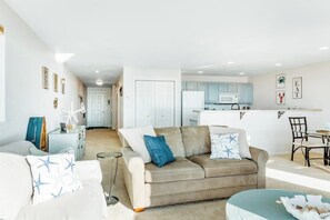 Ein modernes Wohnzimmer mit einem beigen Sofa, einem blauen Pouf und einem weißen Sessel. Im Hintergrund ist eine Küche mit hellblauen Schränken und einem weißen Kühlschrank erkennbar.