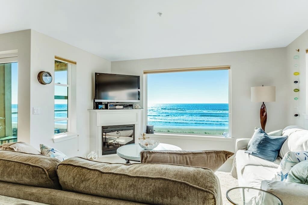 A living room with a large window overlooking the ocean, a flat-screen TV, and a fireplace.