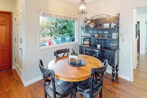 A dining area with a round wooden table and chairs, a window with a view of greenery, and a dark wooden shelf with glassware. - Prime 1BR Oceanfront Dog Friendly - Patio - Washer Dryer (Port Angeles)