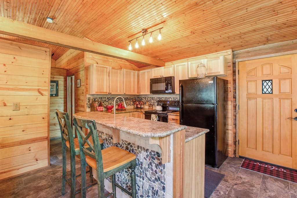 A wooden cabin interior with a kitchen featuring a granite countertop, a black refrigerator, and a stone backsplash.