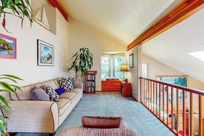 Ein gemütliches Wohnzimmer mit einer Couch, einem Couchtisch und einem Bücherregal. Eine Treppe führt zu einer oberen Ebene mit einer Handläufe.