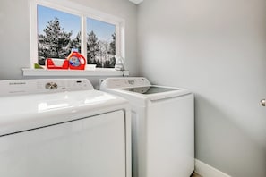 A laundry area with a Samsung washer and dryer, a window with a view of trees, and a shelf with laundry detergent. - Modern 3BR Dog Friendly - Hot Tub - Patio (Westport)